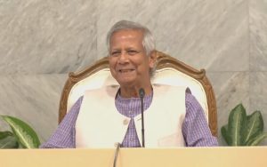 Dr. Muhammad Yunus, a renowned economist and social entrepreneur, smiling and seated in a formal setting. He is wearing a traditional Bengali outfit with a beige vest, and the background features a marble wall with some indoor plants.