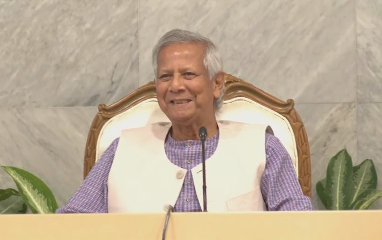 Dr. Muhammad Yunus, a renowned economist and social entrepreneur, smiling and seated in a formal setting. He is wearing a traditional Bengali outfit with a beige vest, and the background features a marble wall with some indoor plants.